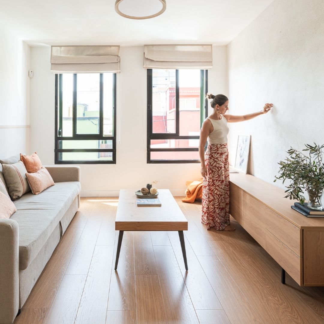Salón luminoso con sofá beige, cojines en tonos tierra y mesa de madera, acompañado de un mueble bajo con planta decorativa y ventanas que dejan entrar abundante luz natural, en un proyecto de interiorismo en Tenerife.
